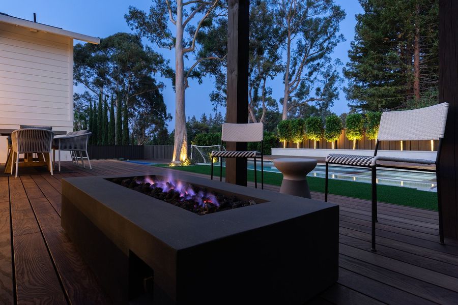 Concrete fire table with a purple flame at dusk, woven chairs and a lit hedge in the background