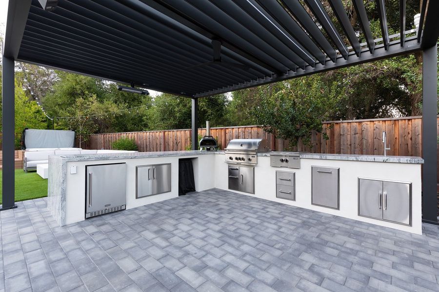 Outdoor kitchen island with a veined stone counter, built-in grill, pizza oven and stainless cabinets under a louvered pergola