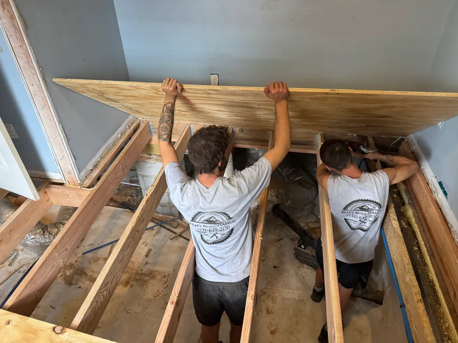 Two Ben’s Busy Hands crew members in company shirts lifting subfloor over new joists